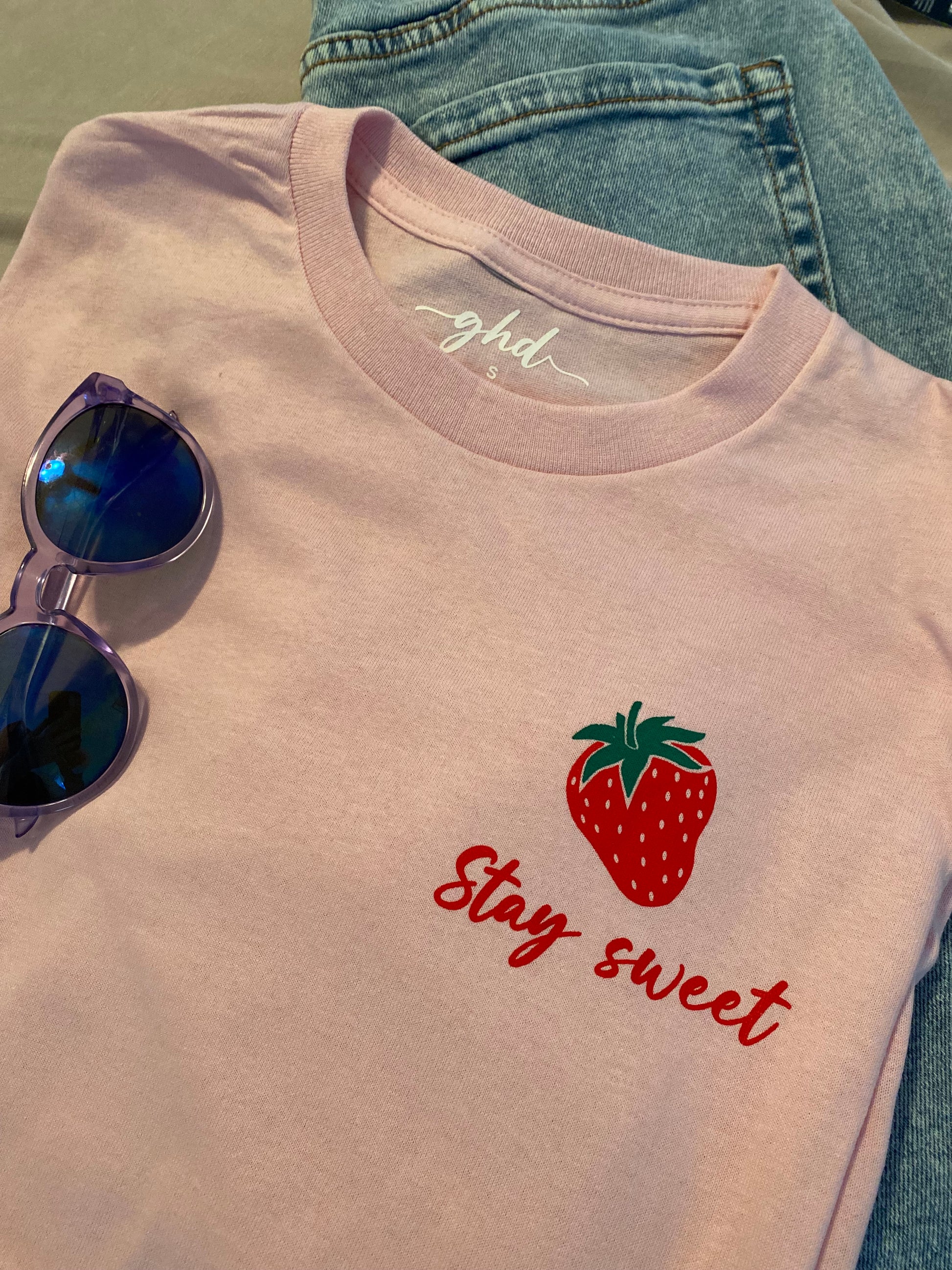 Stay Sweet t-shirt folded on top of a pair of blue jeans, with purple sunglasses laying on top of the tee. The tee is in a light pink colour with red handwriting that says "Stay Sweet" underneath a single red strawberry with a green stem.  The design is located on the upper left side of the tee. 