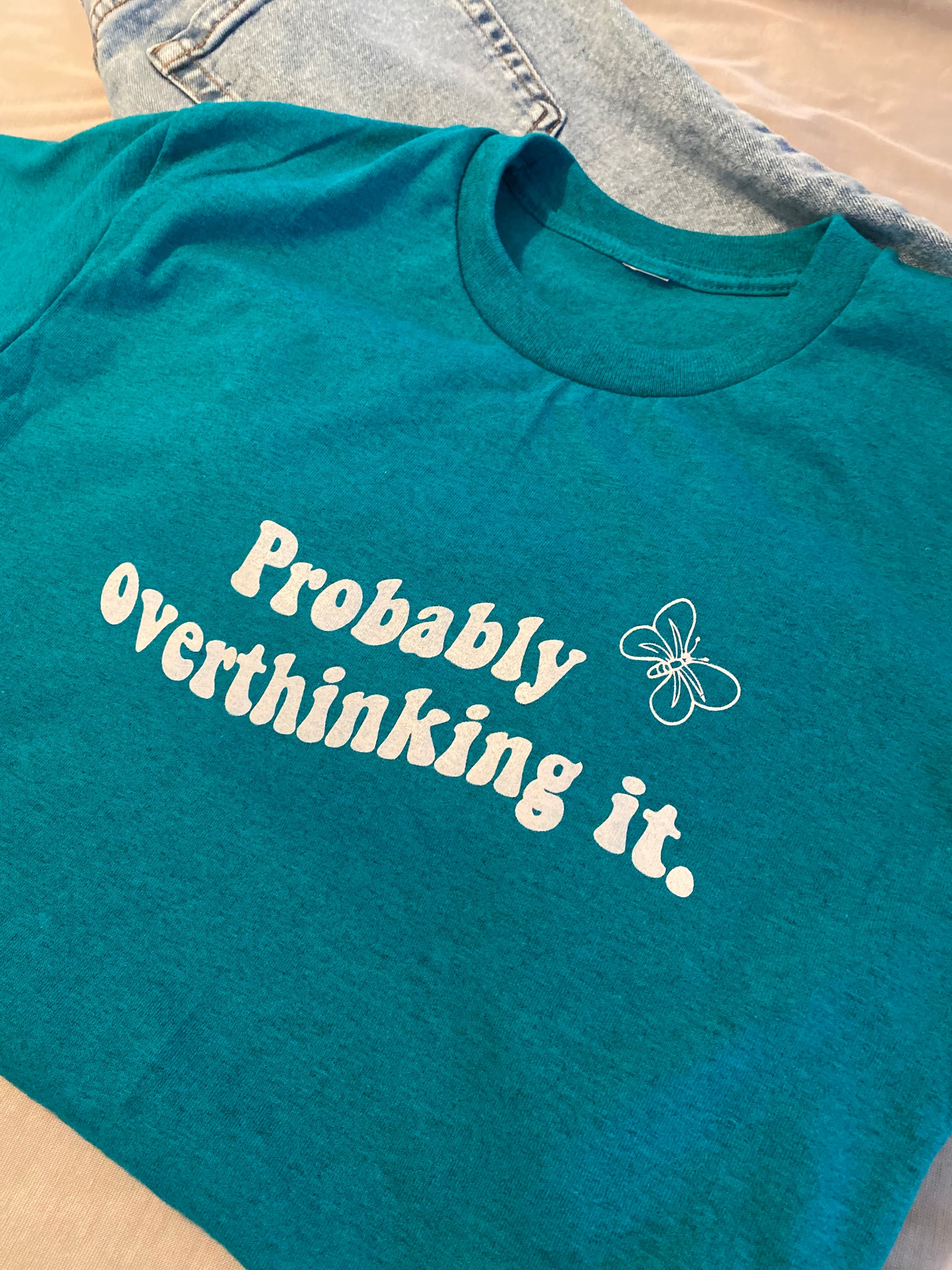 Probably overthinking it tee, in white print with a wave design centered in the middle of the shirt. There is also a white coloured butterfly in the top left corner. colour is in antique sapphire. It is folded on a pair of blue jeans . 