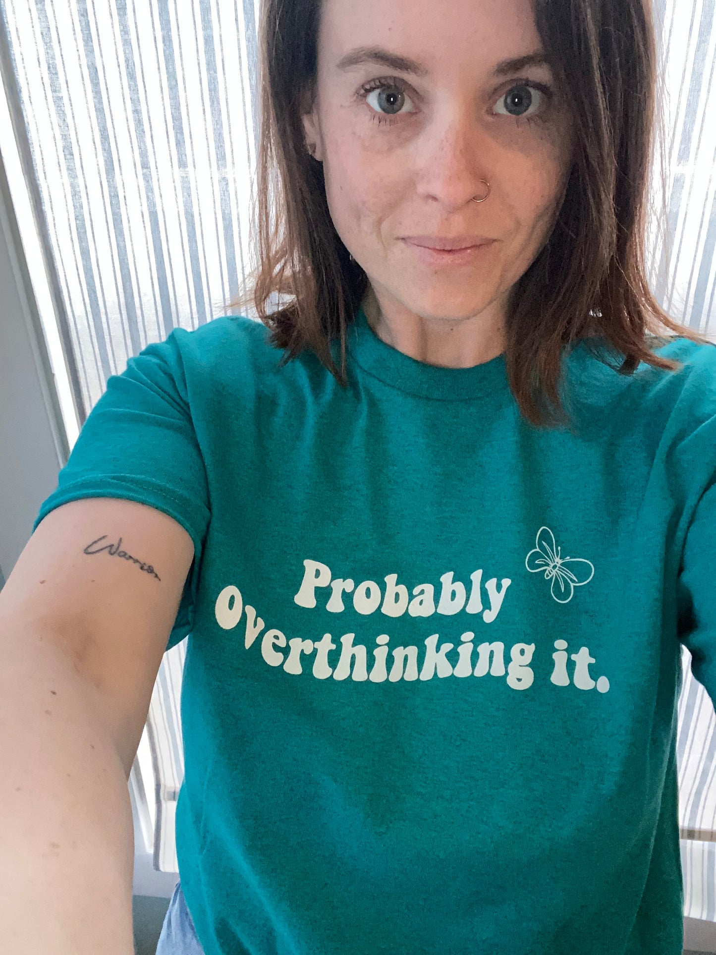 woman taking selfie wearing the Probably Overthinking it tee, from the waist up. The t-shirt writing is in white, with a wave design and a butterfly on the top left corner, also in white. Colour is in antique sapphire. The design is centered in the middle of the t-shirt. The material is light weight and soft, model is wearing a size medium.