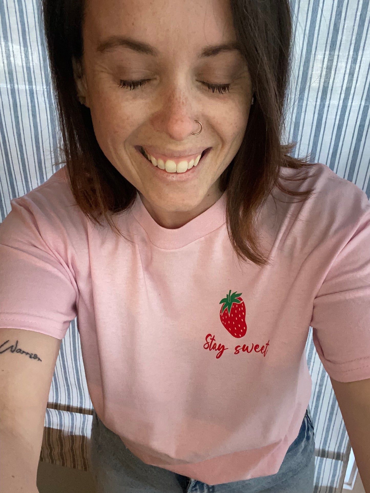 Woman talking a selfie wearing the Stay Sweet tee. The picture is from the waist up and she is smiling with her eyes closed. The tee is tucked into a pair of blue jeans. it is loose fitting the way the model is wearing it. The t-shirt is light pink coloured with red handwriting for the "Stay Sweet " and it is placed underneath a single red strawberry with a green stem, located in the upper left side of the t-shirt. 