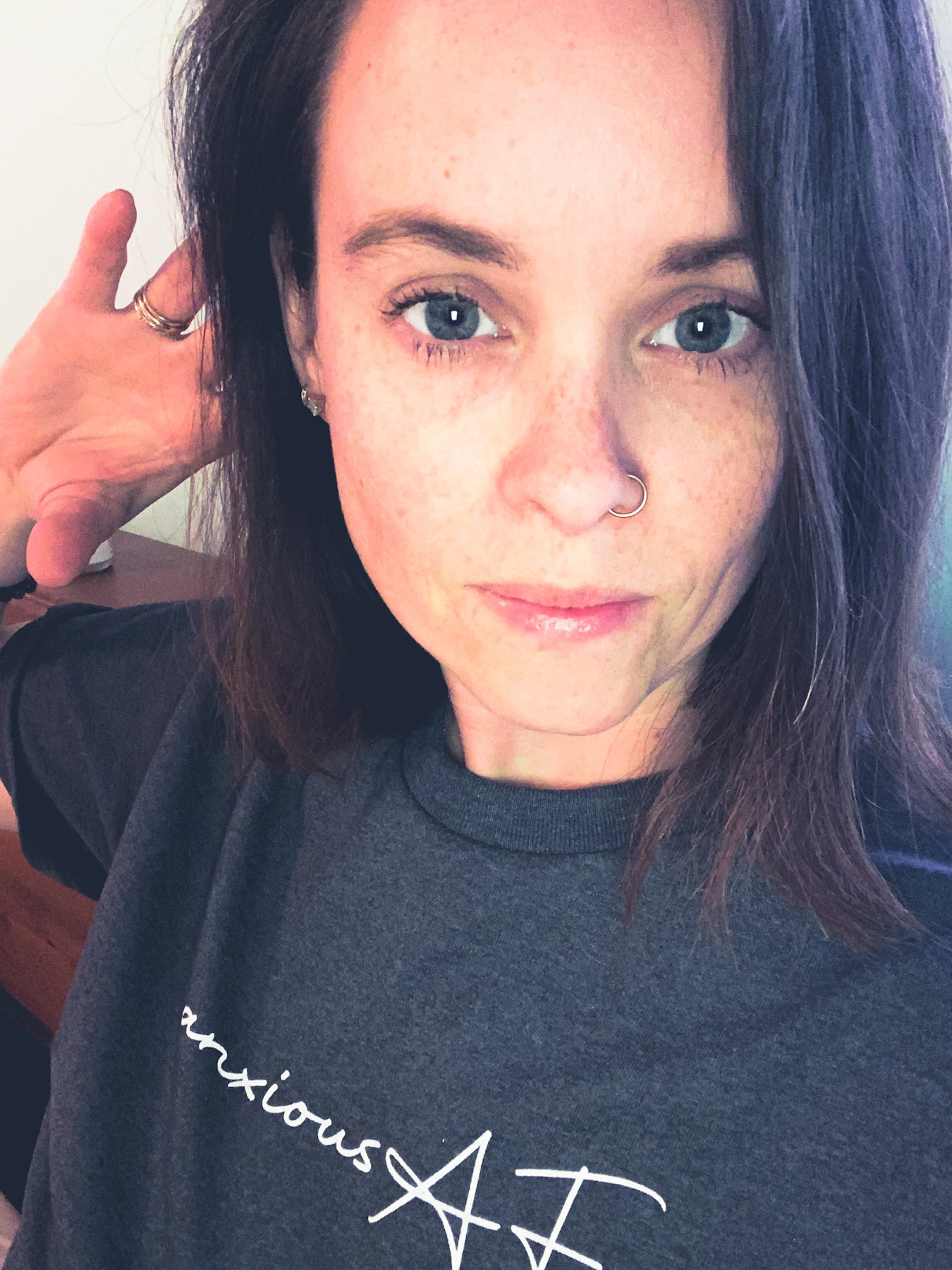 anxious AF tee, in the colour tweed. The design is centered in the upper portion of the t-shirt, with white writing. Anxious is printed in a lower case cursive font and AF is printed in a uppercase cursive font. The model is taking a close up selfie with her right hand placed beside her right ear, arm is bent and facing outwards from the body.  Picture is from just the bottom of the writing and upwards. 