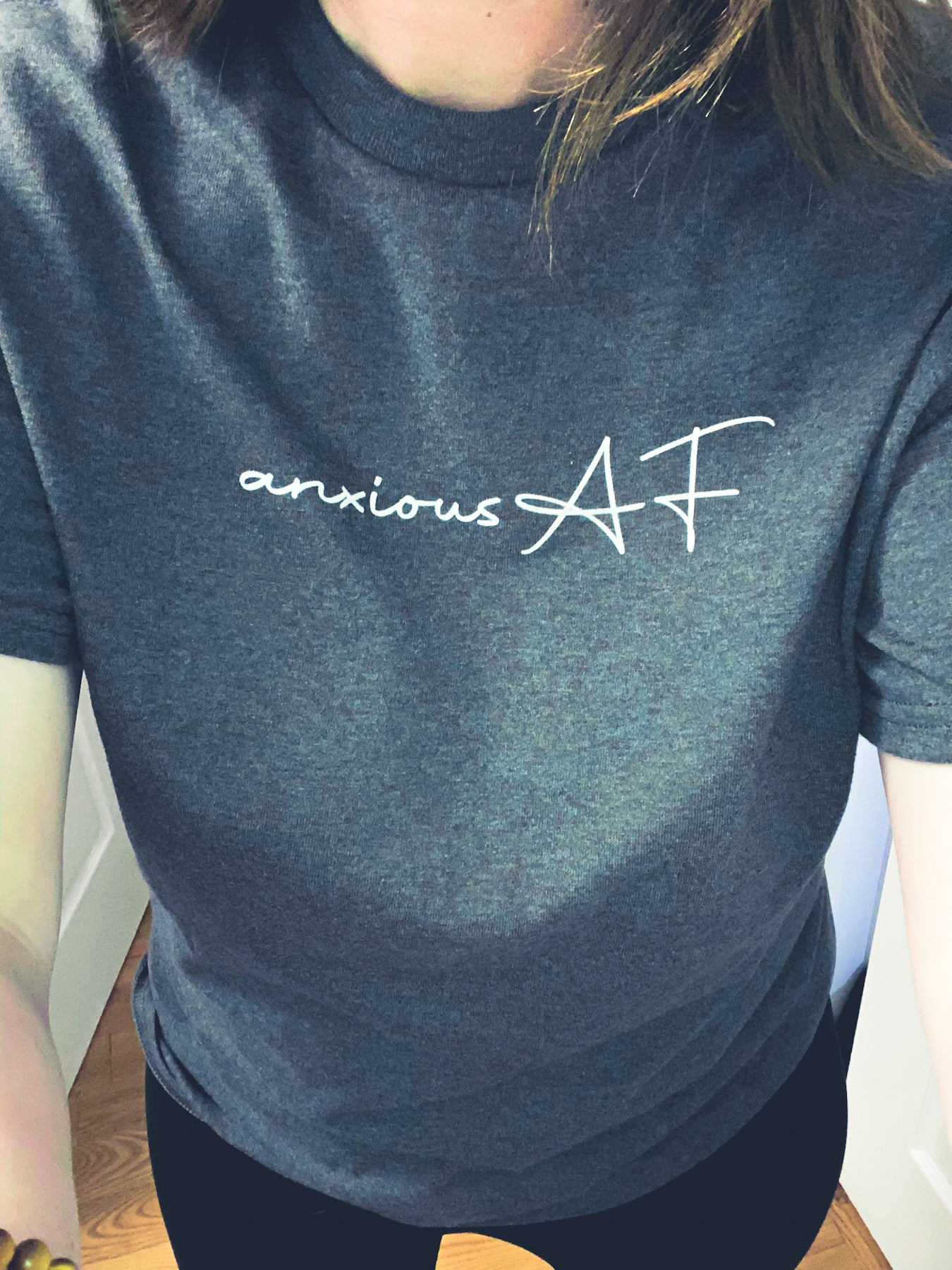 Picture is of anxious AF tee, in a tweed colour. Model is taking a selfie of the middle section of the body. The design is located in the middle upper section of the t-shirt. the writing is in a white coloured print with "anxious" in lower case cursive font and "AF" in a upper case cursive font. 