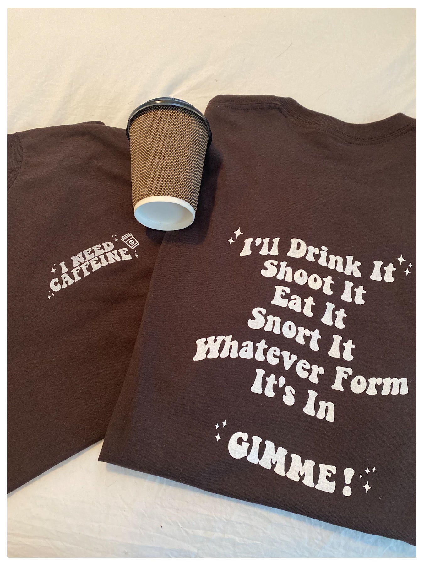 Folded up front and back version of "I NEED CAFFEINE," tees side by side with a brown take out coffee cup on top, in the middle of the two. the colour of the t-shirt is in the colour chocolate, and is light weight. 