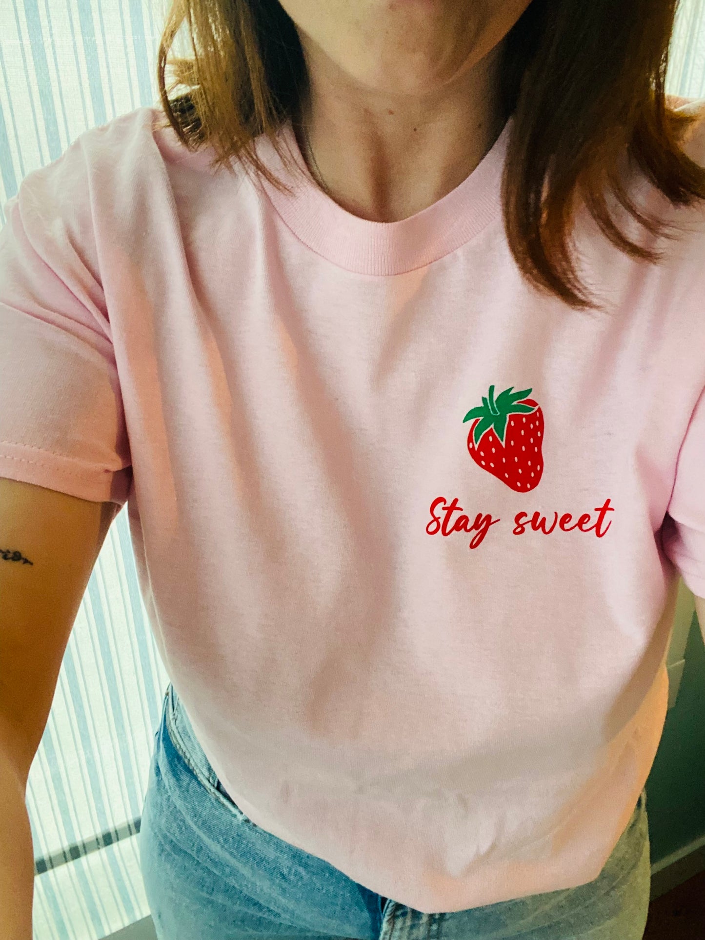 Woman wearing "Stay Sweet" t-shirt. Picture is taken of the mid section of the body. The tee is tucked into a pair of blue jeans, flowing loosely. The t-shirt is light pink coloured with a soft material. The "Stay Sweet" is written in red ink and a cursive font. It is centered underneath a single red strawberry with a green stem. The design is located in the upper left side of the t-shirt.  