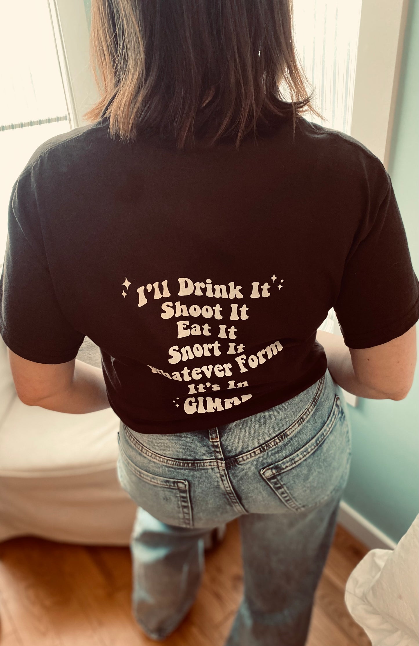 Back view of the 'I NEED CAFFEINE" tee. It says "I'll Drink It, Shoot It, Eat It, Snort It, Whatever Form, It's In, GIMME!" The writing is white ink as well, located in the middle back of the tee. The t-shirt is in the colour chocolate.  