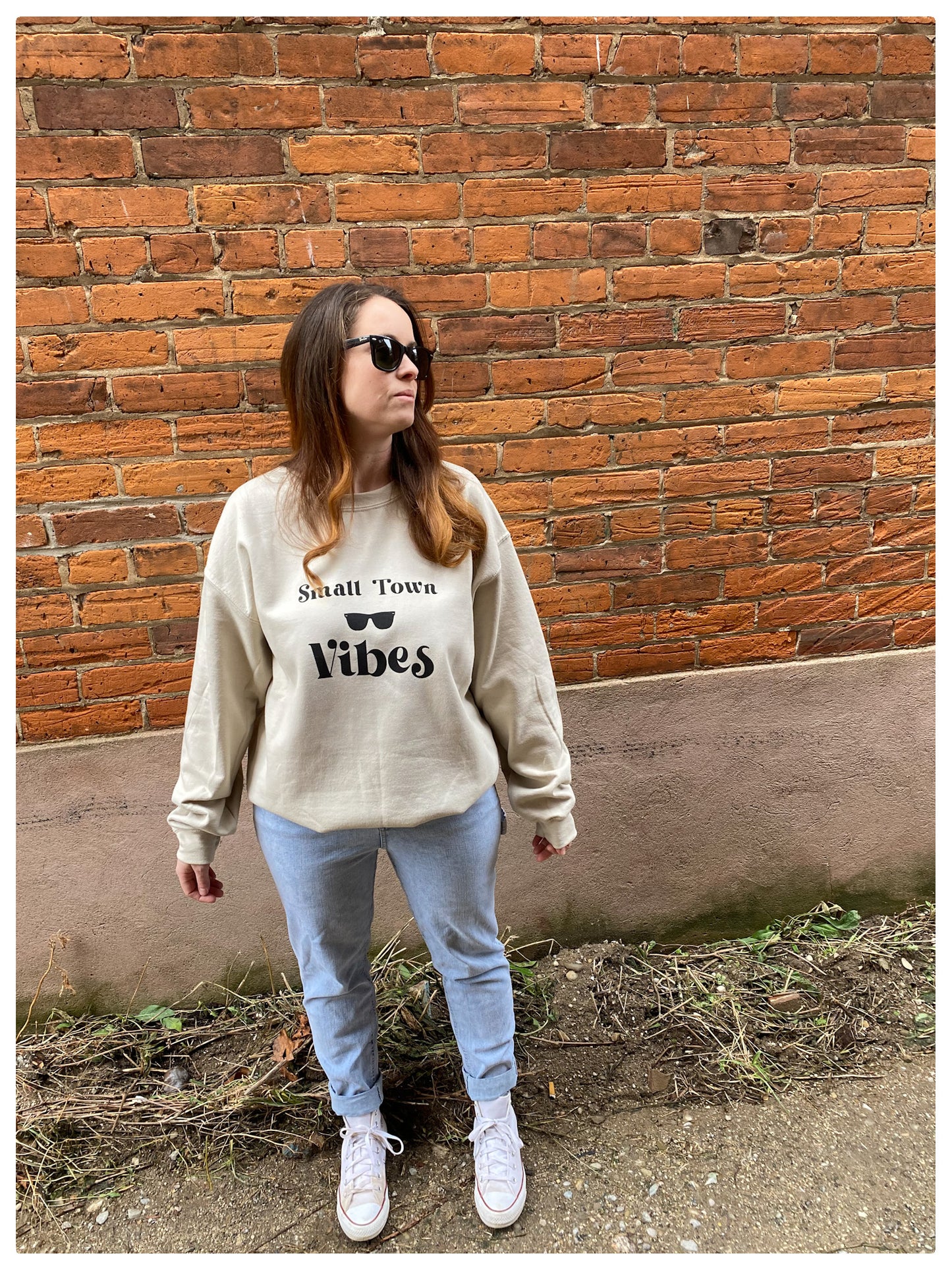 Girl standing against a brick wall wearing a warm  sand colour sweater that says "Small town vibes"