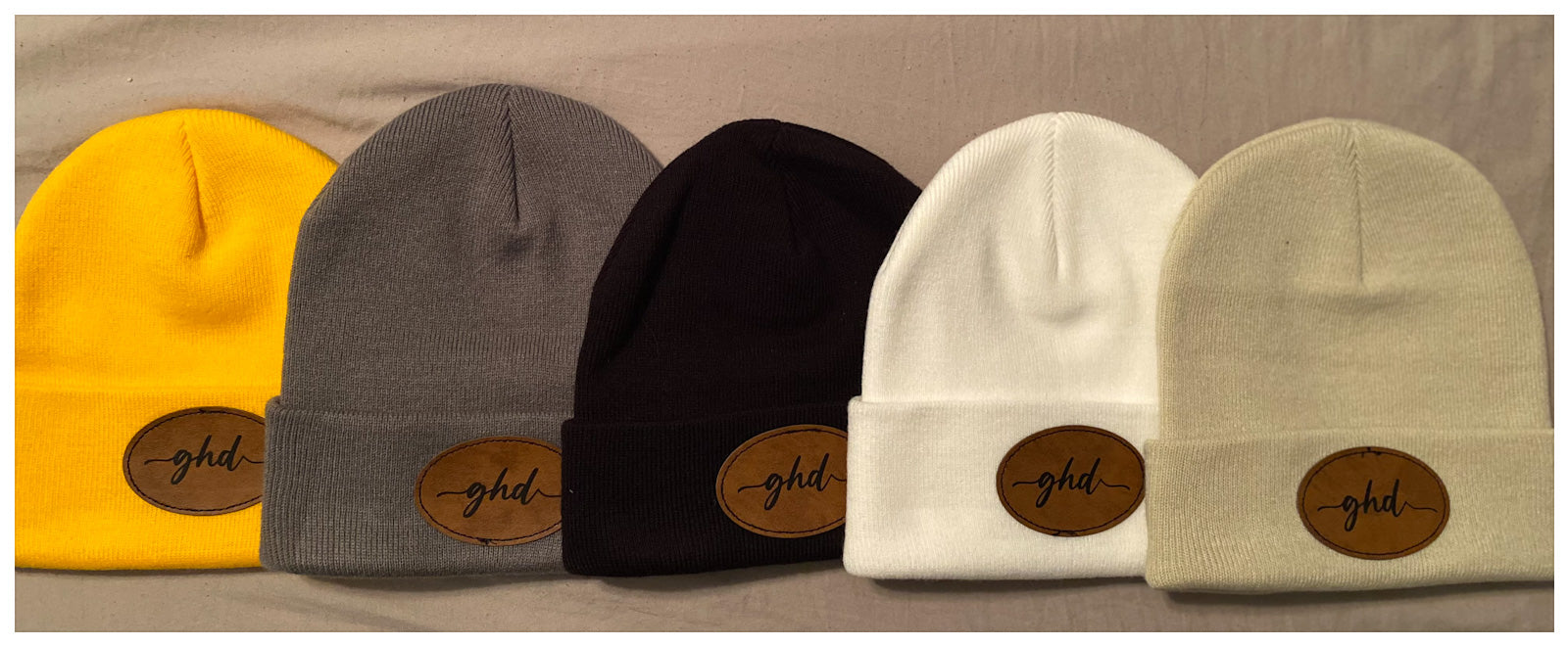 Winter beanies in 5 different colours laying side by side with "ghd" written on a brown faux leather patch in black. Colours; gold, grey, black, white and ivory.