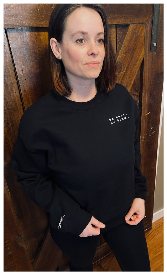 Woman standing in front of a barnboard door, she is wearing a cozy black fleece crew that says "be cool, be kind." written on the upper left side. 