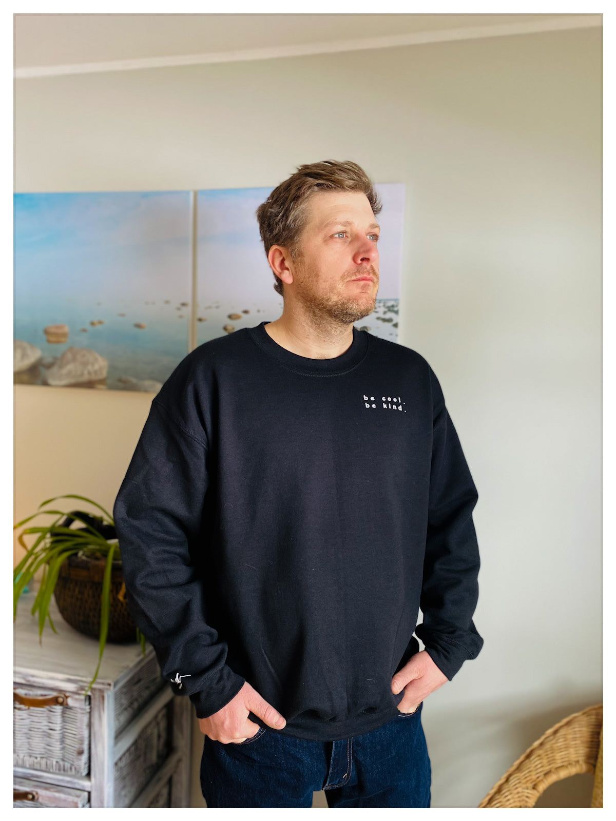 Man standing with hands in his pockets . He is wearing a cozy fleece black crew that says "be cool, be kind" written on the upper left side. 