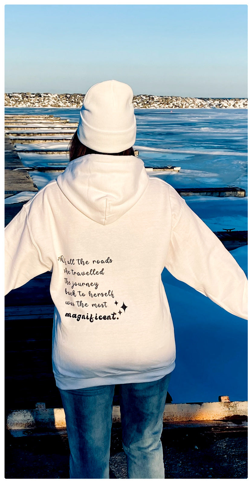 A woman standing in front of a harbour with her back to the camera, she is wearing a white beanie and a white cozy fleeced hoodie that says written on the left side in black cursive writting. "Of all the roads she travelled the journey back to herself was the most magnificent." Her arms are stretched out at both of her sides. 