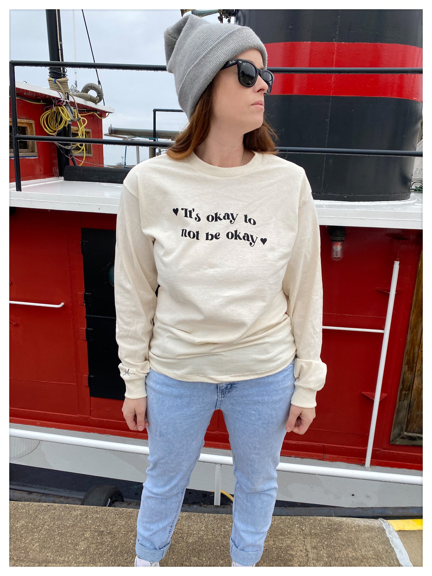 girl standing in front of boat wearing a natural coloured long sleeved tee that says "It's Ok To Not Be Ok "