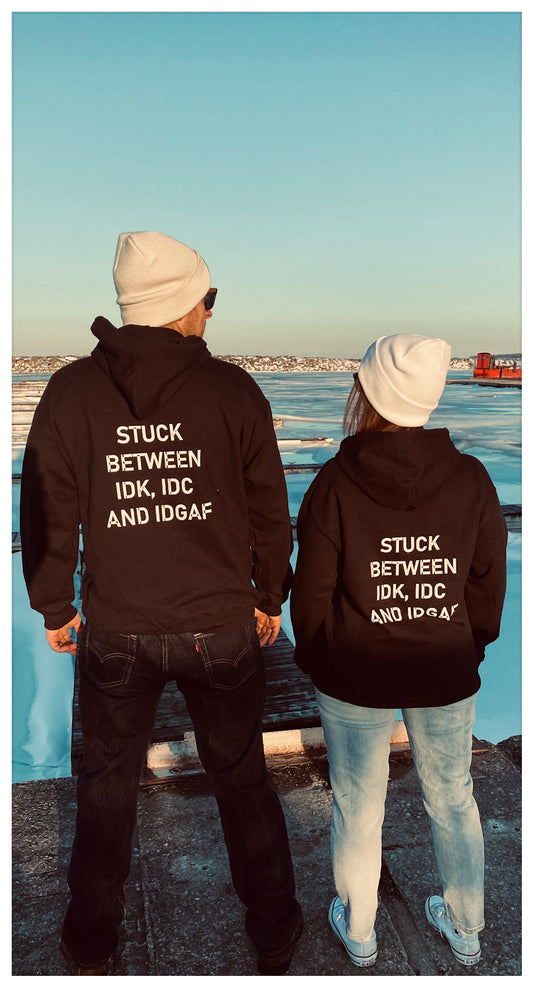 A man and a woman with their backs facing the camera wearing the same black hoodie that says '" stuck between IDK, IDC, AND IDGAF.  They are both also wearing ghd winter beanies and jeans.