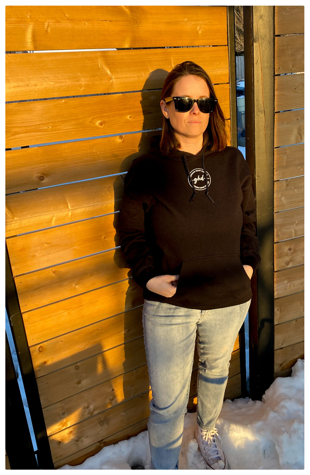 Woman standing in front of a wooden gate wearing the black Stuck between IDK,IDC, AND IDGAF hoodie. with the ghd logo printed in white print on the front center of the sweater.  She is also wearing, jeans, black sunglasses and white sneakers. 