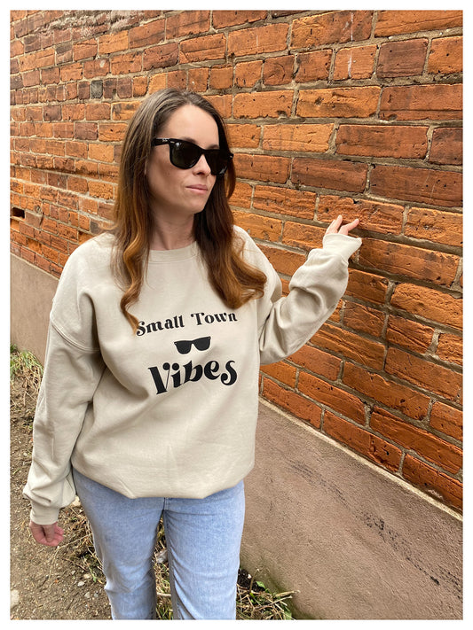 Girl standing against a brick wall with a cozy  sand coloured crew that says "small town vibes"