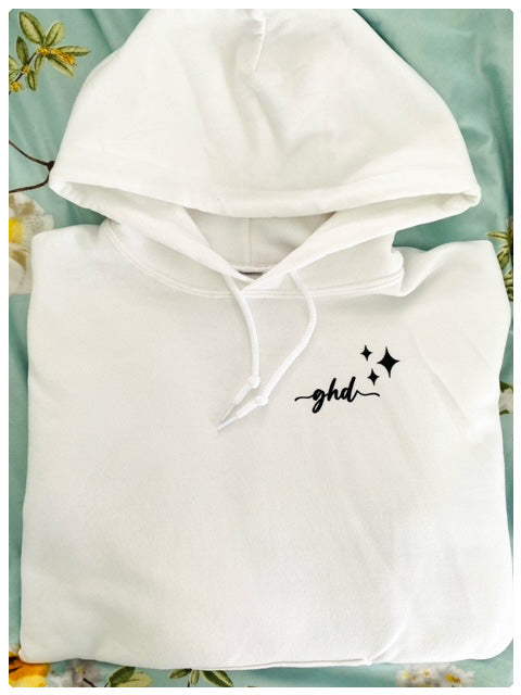 Folded white hoodie on a flowered blanket that has the "ghd" logo printed in black cursive writing on the upper left side with three different sized stars beside it.  